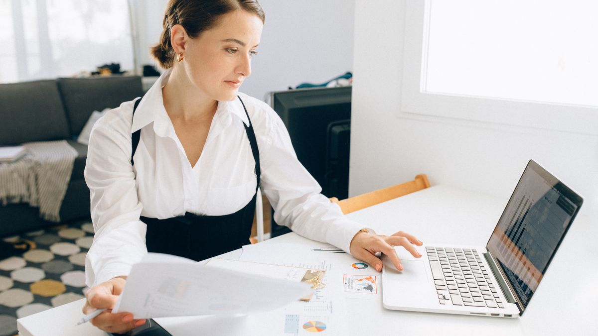 Woman at desk reviewing business reports on laptop, holding papers in a modern office setting.